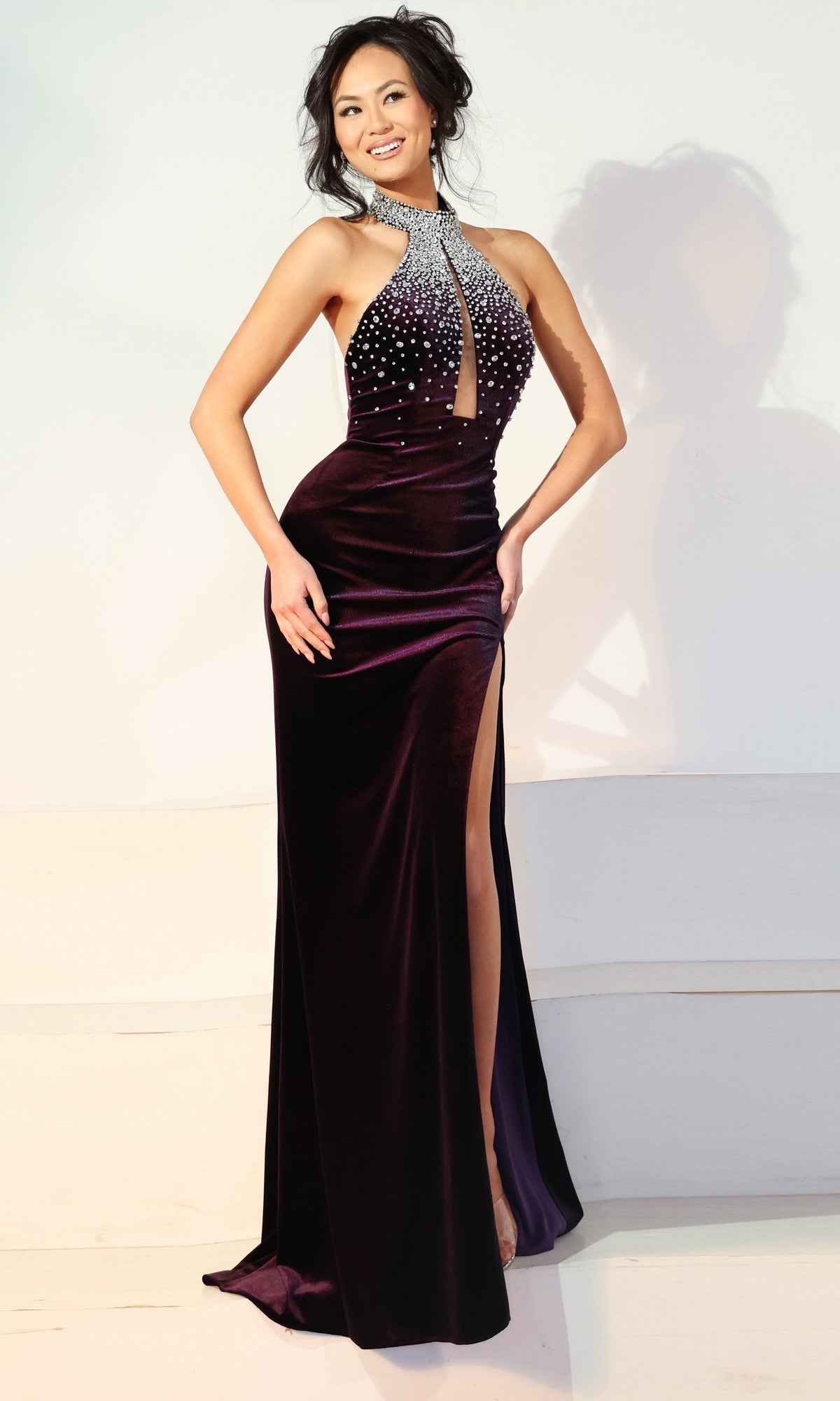 A woman stands smiling in the Jovani 48960 Long Prom Dress, featuring a dark purple hue, crystal embellishments, a high neckline, pearl accents, and a high leg slit.